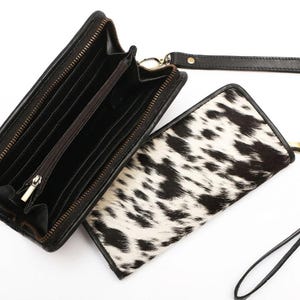 May include: A black and white cowhide print wallet with a wrist strap. The wallet is open, revealing a black interior with multiple card slots, a central zippered compartment, and a full-length bill pocket. The exterior features a textured fur pattern.