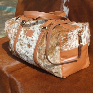 May include: A brown leather duffel bag with a white and brown cowhide pattern. The bag has a top handle and a shoulder strap.