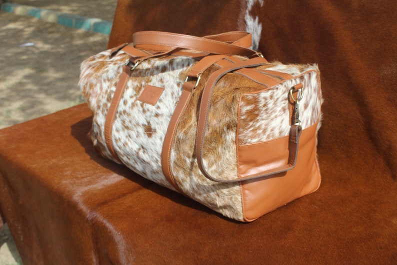 May include: A brown leather duffel bag with a white and brown cowhide pattern. The bag has a top handle and a shoulder strap.