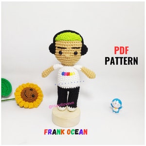 Può includere: Schema di amigurumi all'uncinetto per una bambola Frank Ocean. La bambola indossa una maglietta bianca con un logo arcobaleno, pantaloni neri e cuffie. Lo schema è disponibile per il download in PDF.
