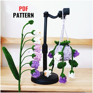 Op de afbeelding: Een gehaakt bloemenboeket met paarse en witte bloemen hangend aan een paarse gehaakte pot. De pot hangt aan een zwarte standaard. Het boeket staat op een houten oppervlak. De tekst "PDF PATTERN" staat in rode letters bovenaan de afbeelding. De tekst "@handlovevn" staat onderaan de afbeelding.