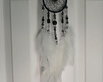 May include: A handcrafted dreamcatcher with a black hoop and intricate web design. The dreamcatcher features black and clear beads, and white feathers. It hangs from a black leather strap, creating a decorative wall hanging.