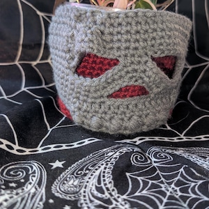 May include: A gray crocheted plant pot cover featuring a face with two angular eye cutouts and a horizontal mouth cutout, revealing a dark red inner layer. The cover is cylindrical and textured with visible yarn stitches throughout the exterior.