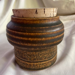 May include: A brown ceramic jar with a cork lid. The jar has a textured surface and a striped pattern.