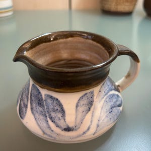 May include: A small ceramic creamer with a rounded body and a dark brown rim. The creamer has a white base with blue leaf-like patterns. It features a small handle and a spout for pouring.