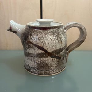 May include: A ceramic teapot with a lid and handle. The teapot is mainly brown with white and dark brown streaks and a white spout. The lid is white with a dark red rim. The handle is a curved shape.