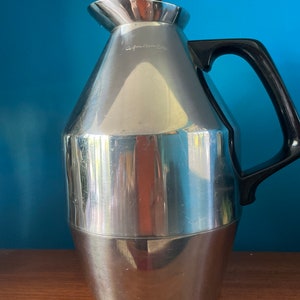 May include: A silver stainless steel thermal carafe with a black handle and a black lid. The carafe has a rounded body and a slightly flared spout.