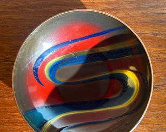 May include: A small, round decorative bowl with a dark rim and a colorful interior. The bowl's interior features a vibrant design of red, blue, and yellow curved lines against a dark background. The bowl sits on a wooden surface.
