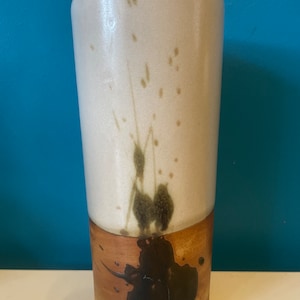 May include: A white ceramic vase with brown and green splattered paint. The vase has a cylindrical shape and a brown base.