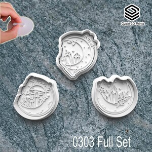 May include: Three white plastic cookie cutters with nature-inspired designs. One features a crescent moon with crystals, another has mushrooms, and the third has crystals and flowers. The text "0303 Full Set" is at the bottom. The cutters are on a grey surface.