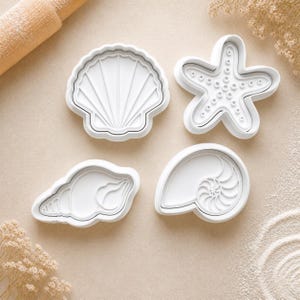 May include: A set of four white plastic cookie cutters in the shapes of a scallop shell, a starfish, a conch shell, and a nautilus shell. Each cutter features detailed embossed lines to create impressions in dough or clay for baking or crafting.
