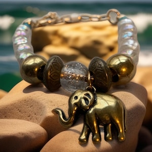 May include: A beaded bracelet featuring a gold elephant charm. The bracelet includes gold, clear, and patterned beads. The elephant charm is detailed with a trunk and tusks. The bracelet is displayed on a sandy surface with a blurred ocean background.