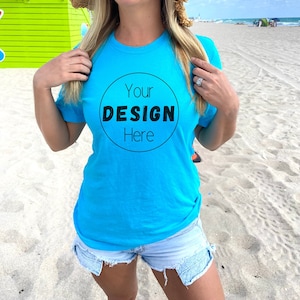 Puede incluir: Una mujer con una camiseta azul turquesa con un círculo negro en el frente que dice "Your Design Here" y un sombrero de paja. Está de pie en una playa de arena.