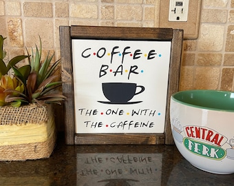 May include: A coffee-themed display featuring a framed sign with "COFFEE BAR" and a coffee cup graphic, along with a large "Central Perk" mug and a small potted plant. The sign reads "The One With The Caffeine."