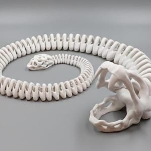 May include: A white ceramic snake sculpture, coiled in a spiral, with a detailed head and body segments. The sculpture is on a grey surface. The snake's mouth is open, revealing its teeth.