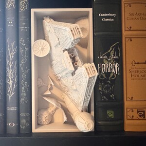 May include: A white spaceship model is presented in a white box, with classic book spines in the background. The books include titles such as "Classic Tales of Horror" and "Sherlock Holmes".