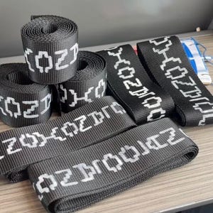Peut inclure: Plusieurs rouleaux et longueurs de sangle noire avec du texte blanc qui dit "(OZ)" et "OZPRODUCT". La sangle est présentée sur une surface brun clair. Les rouleaux sont bien enroulés et les longueurs sont posées à plat.