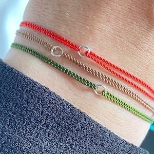 May include: Three bracelets on a wrist. The bracelets are red, green, and brown cord with silver rings.