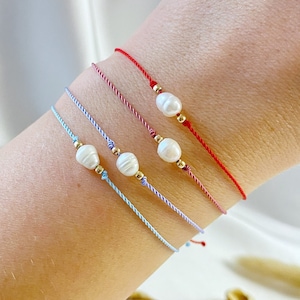 Puede incluir: Cuatro pulseras con cuentas de perlas y detalles dorados en una muñeca. Las pulseras son rojas, rosadas, azul claro y azul oscuro.