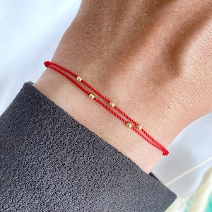 May include: Red string bracelet with gold beads. The bracelet is tied on a person's wrist.