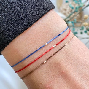 May include: Three bracelets on a wrist. The bracelets are made of thin cord in blue, red, and brown. Each bracelet has a small silver bead.