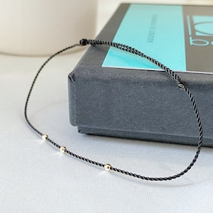 Pode incluir: Um bracelete de corda preta com três pequenas contas douradas. O bracelete está sobre uma superfície branca e uma caixa preta com o texto "barandi 14K GOLD" está no fundo.