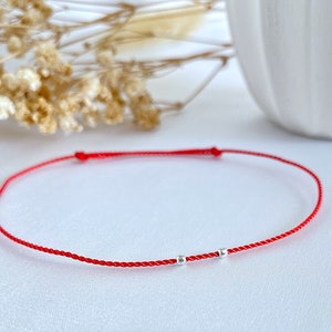 Op de afbeelding: Een rood koord armbandje met twee kleine zilveren kralen. Het armbandje ligt op een wit oppervlak.
