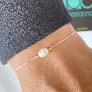 May include: A delicate bracelet featuring a single white pearl and gold accents on a pink string.