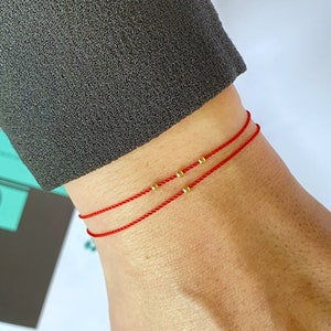 May include: Two red string bracelets with small gold beads on a person's wrist.