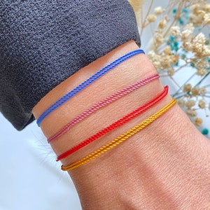 Könnte beinhalten: Vier farbige Armbänder, die auf dem Handgelenk einer Person übereinander liegen. Die Armbänder sind blau, rosa, rot und gold.