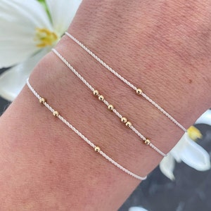 Könnte beinhalten: Drei weiße Armbänder aus Schnur mit goldenen Perlen. Die Armbänder sind übereinander am Handgelenk einer Person gelegt.