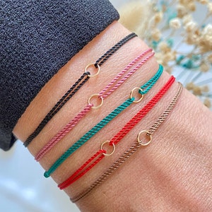May include: A set of six colorful string bracelets with gold rings. The bracelets are layered on a person's wrist, with the black bracelet on top and the brown bracelet on the bottom.
