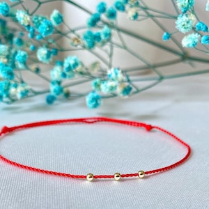 May include: A red string bracelet with three small gold beads. The bracelet is on a white surface.