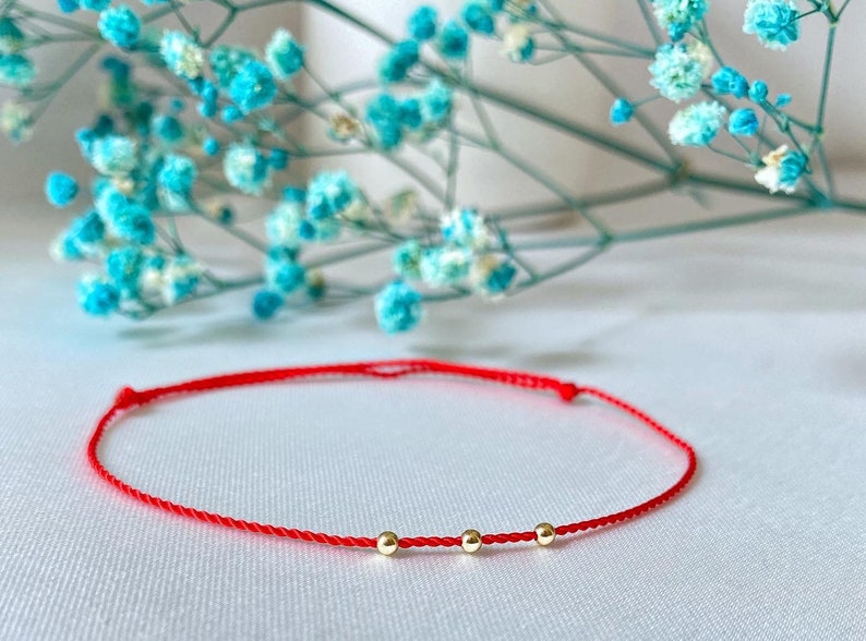 May include: A red string bracelet with three small gold beads. The bracelet is on a white surface.