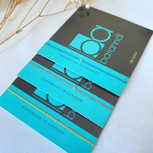 May include: Three colorful string bracelets with a small gold bead on each. The bracelets are stacked on top of each other on a black and turquoise card with the text "Handmade in London" and "14k Gold".
