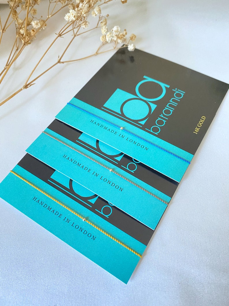 May include: Three colorful string bracelets with a small gold bead on each. The bracelets are stacked on top of each other on a black and turquoise card with the text "Handmade in London" and "14k Gold".
