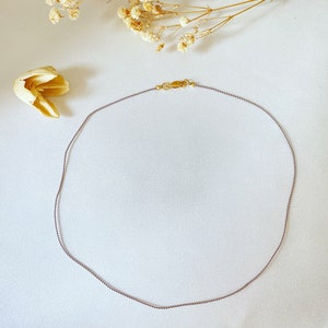 Può includere: Una collana delicata, sottile, in cordoncino viola e bianco con una chiusura dorata.