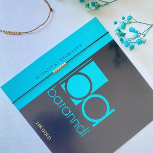 May include: A black string bracelet with gold beads and the text "baramdi" and "14K GOLD" on a turquoise and black background. The bracelet is handmade in London.