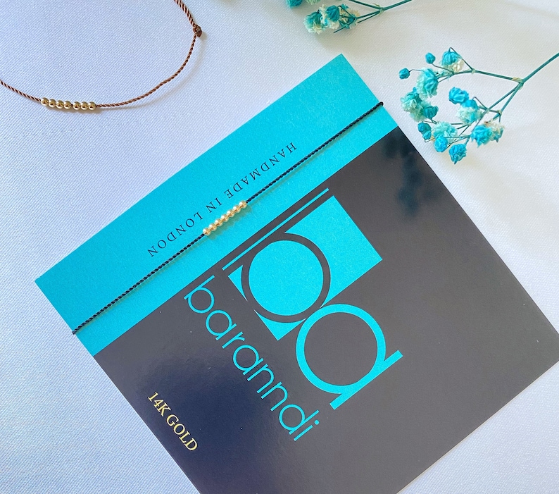 May include: A black string bracelet with gold beads and the text "baramdi" and "14K GOLD" on a turquoise and black background. The bracelet is handmade in London.