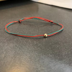 May include: A delicate bracelet with a red and green cord and a small gold bead. The bracelet is a simple design, suitable for everyday wear. The cord is knotted at each end.