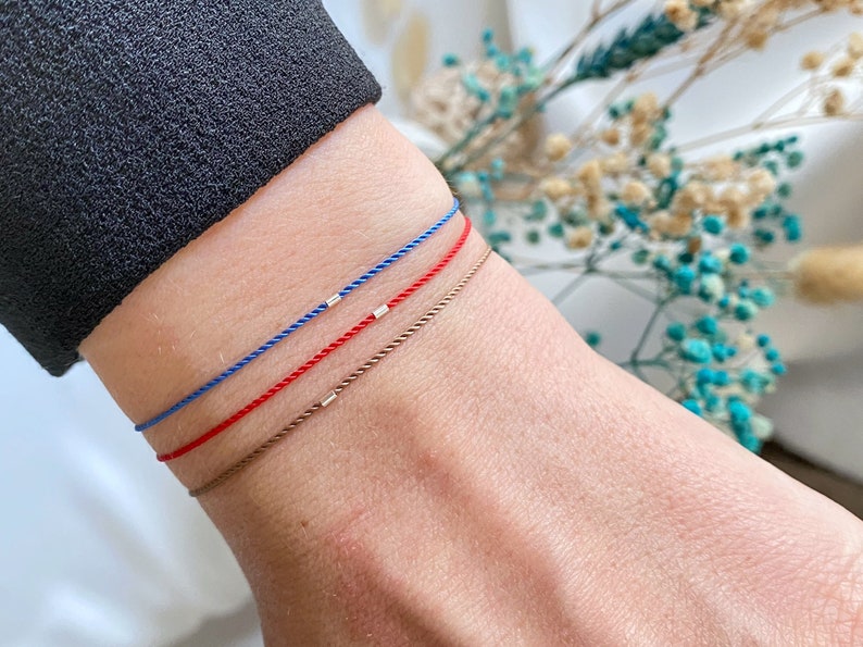 May include: Three bracelets on a wrist. The bracelets are made of thin cord in blue, red, and brown. Each bracelet has a small silver bead.