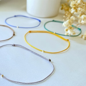 May include: Five colorful string bracelets with a small gold bead accent. The bracelets are arranged on a white surface.