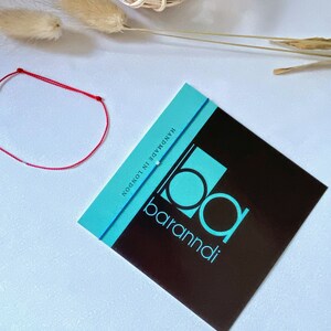 May include: A red and blue string bracelet with white beads. The bracelet is handmade in London by baramndi.