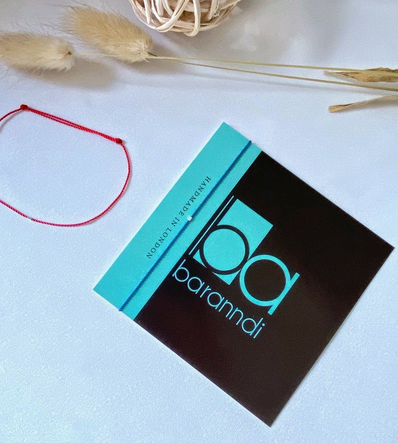 May include: A red and blue string bracelet with white beads. The bracelet is handmade in London by baramndi.