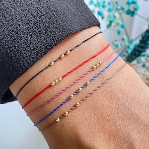 May include: A set of six bracelets with different colored cords and gold beads. The bracelets are stacked on a person's wrist.