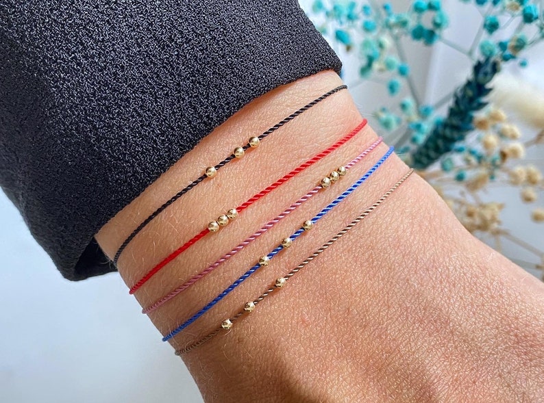 May include: A set of six bracelets with different colored cords and gold beads. The bracelets are stacked on a person's wrist.
