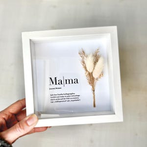 Könnte beinhalten: Ein weiß gerahmtes Kunstwerk mit dem Wort "Mama" in schwarzer Schrift, einer Definition und einem kleinen Strauß getrockneter Blumen. Das Kunstwerk wird von einer Hand gehalten.