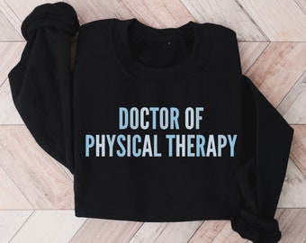 May include: Black sweatshirt with the text "DOCTOR OF PHYSICAL THERAPY" in a gradient blue color. The sweatshirt is displayed on a light brown wooden surface.