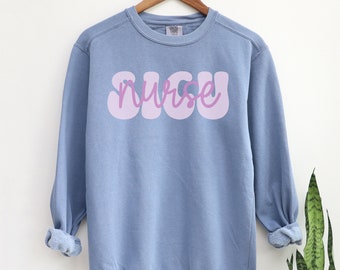 May include: A blue crew neck sweatshirt with the word "nurse" in a bubble font across the chest. The word is in a light purple color. The sweatshirt is hanging on a wooden hanger.