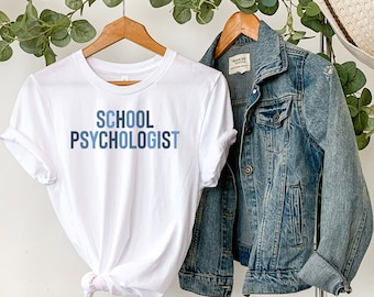 May include: White t-shirt with the words "SCHOOL PSYCHOLOGIST" in blue lettering. A denim jacket and a straw hat hang on a clothing rack. Greenery is draped across the top of the rack.
